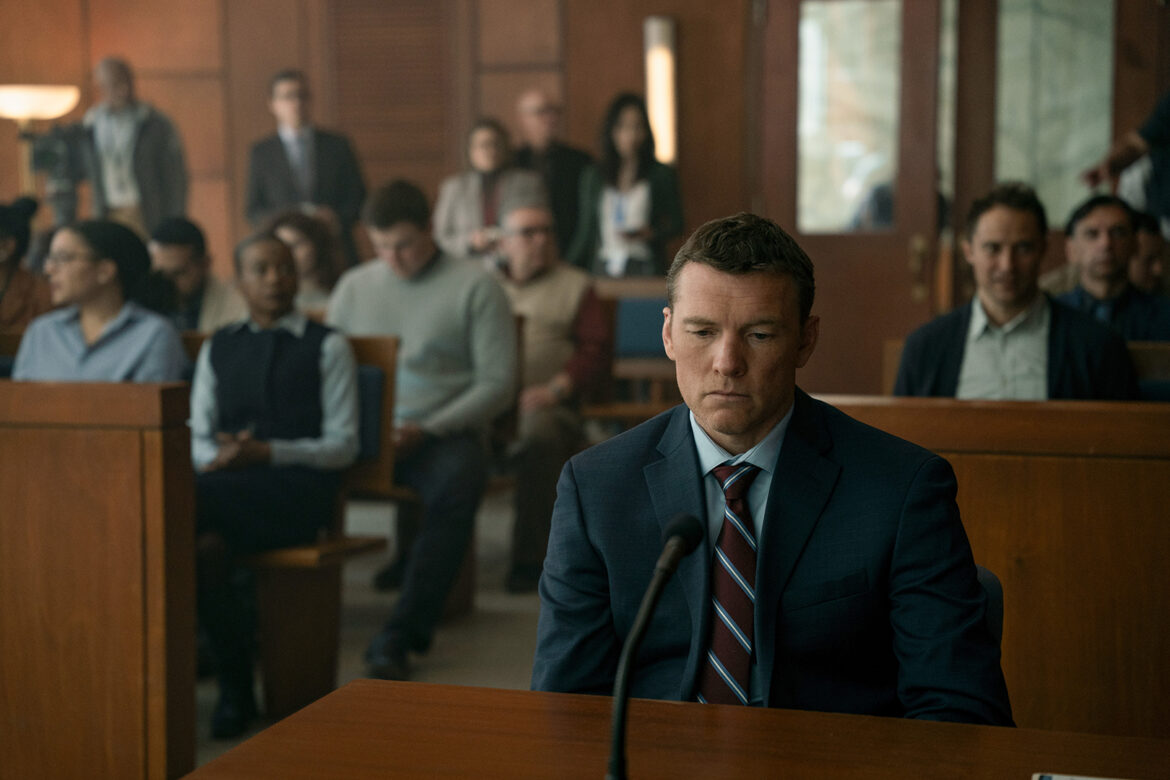 "A man in a suit sits alone at a defendant's table in a courtroom, looking serious. Behind him, an audience of various people watches attentively from the wooden benches, suggesting a tense legal proceeding."